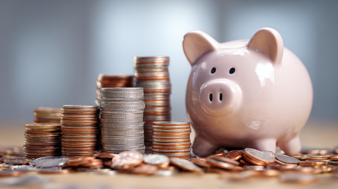 Roze kasica-prasica stoji pored naslaganih kovanica različitih veličina i vrednosti